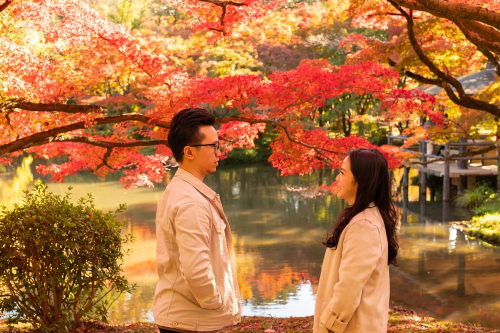 Couple in beige coats standing face to face beside a calm pond surrounded by vibrant red autumn maple leaves in a Japanese garden.