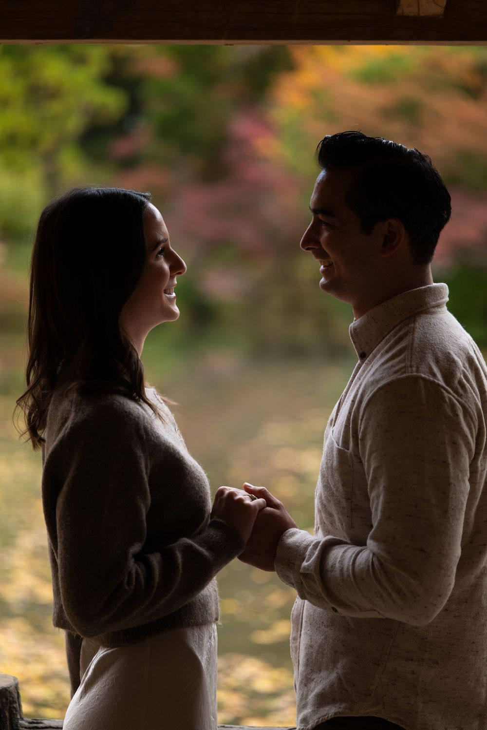 Silhouetted couple holding hands and smiling at each other under a covered area, with colorful autumn foliage glowing in the background.