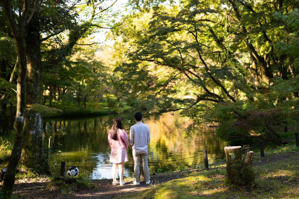 Couple standing side by side on a grassy bank overlooking a calm pond surrounded by lush green trees in warm golden sunlight.