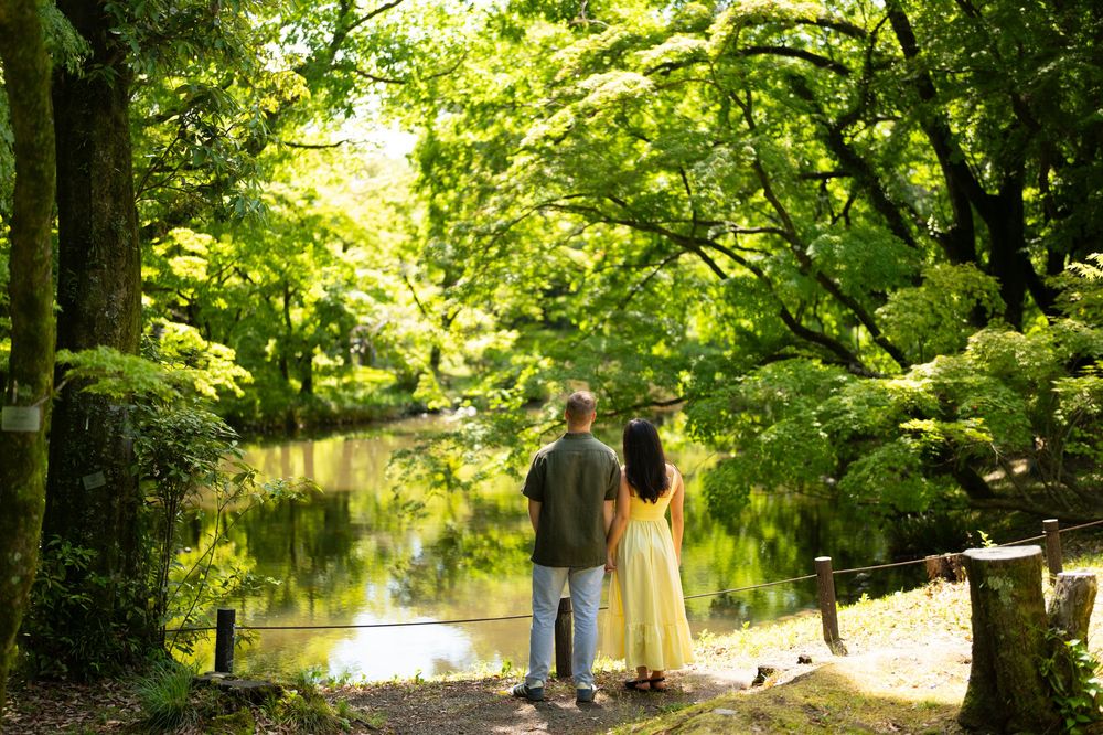 Couple stands side by side on a path beside a calm pond, surrounded by lush green trees in a sunlit park.