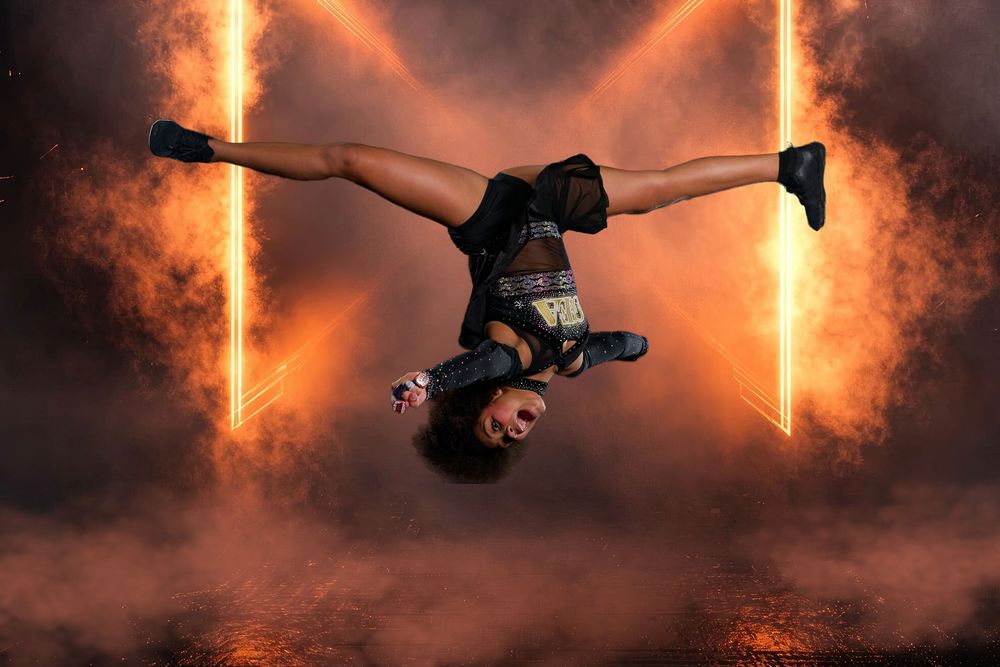 A dancer performs a dramatic backflip mid-air surrounded by glowing orange light beams and smoke.