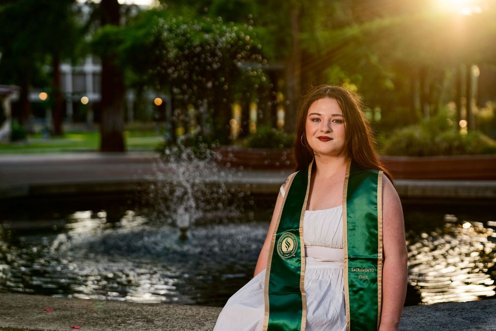 A young woman in graduation regalia poses by a fountain at golden hour on a college campus.