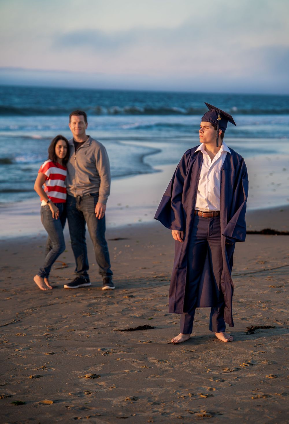 A graduate in cap and gown stands on a sandy beach at sunset with proud parents behind him.