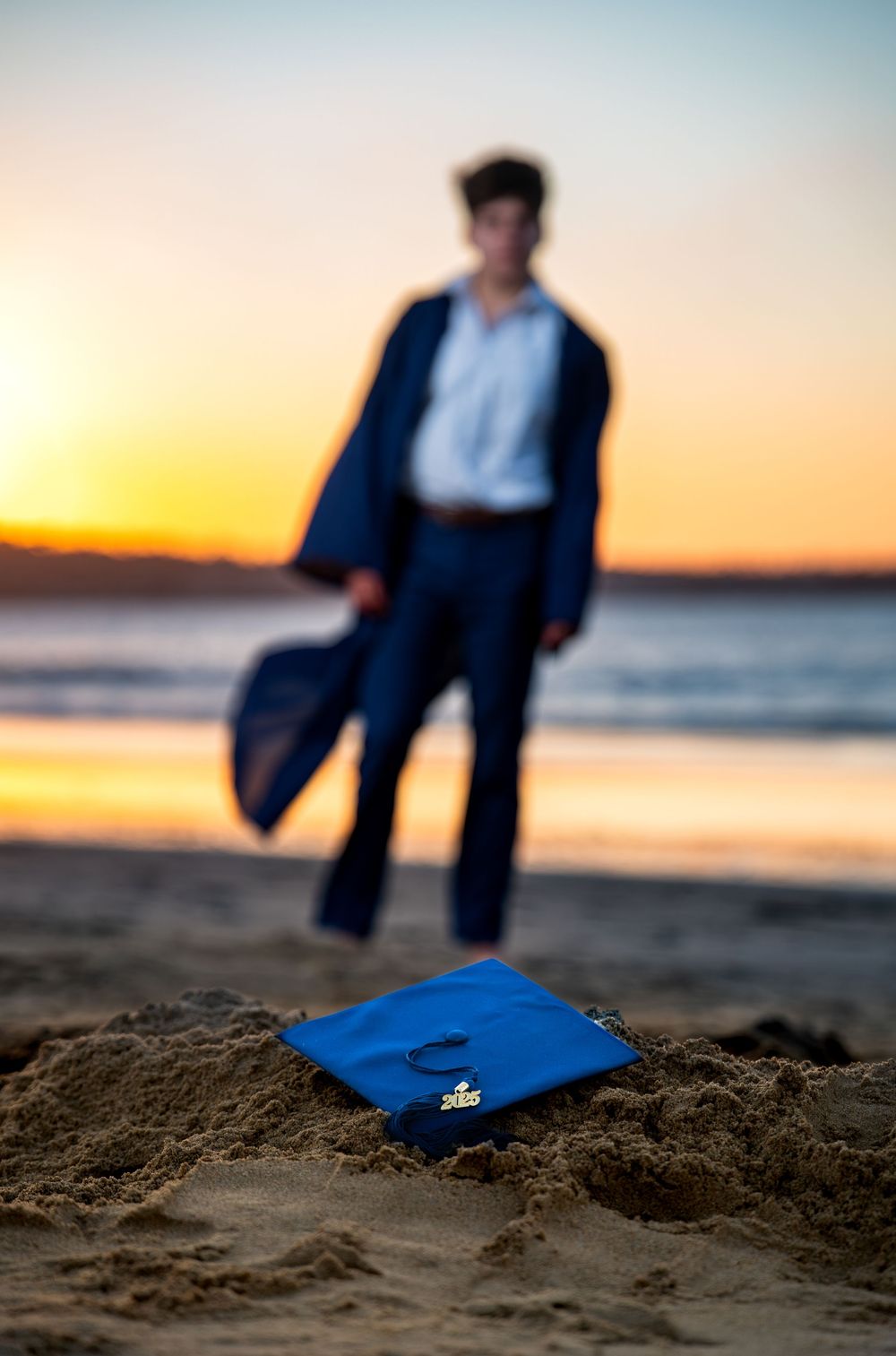 A blue graduation cap rests in beach sand at sunset with a graduate standing in the background.