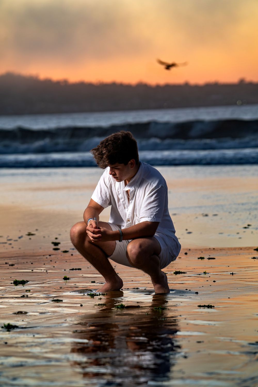 A young man in white crouches on a wet beach during a vivid orange and red sunset.