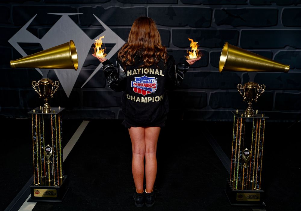 A National Champion dancer stands with her back to camera between two flaming trophies on stage.