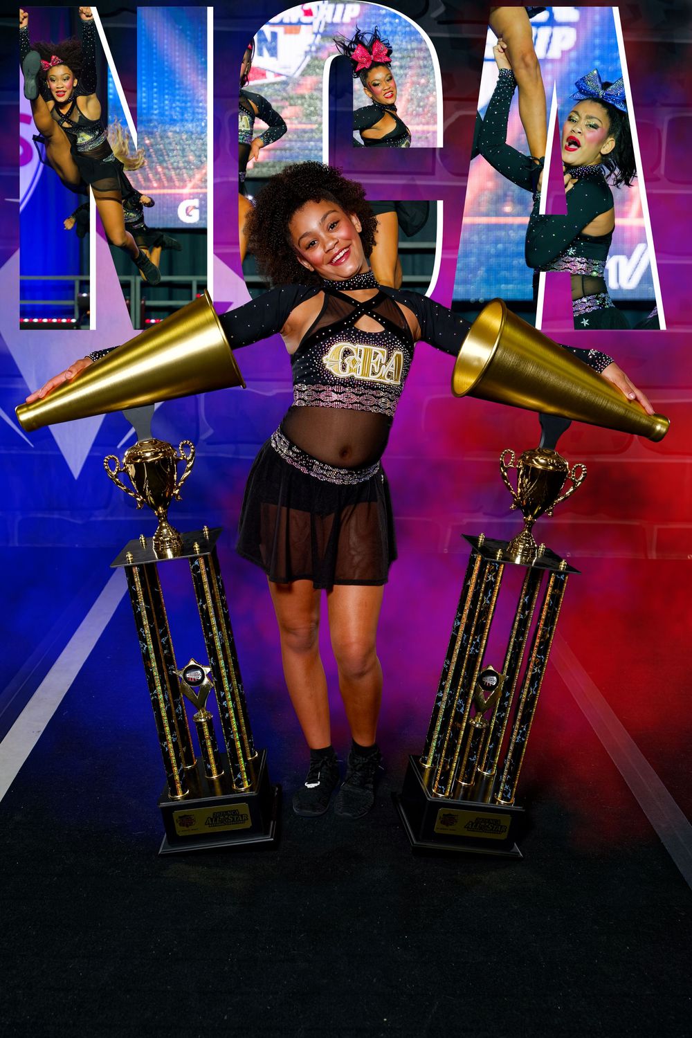 Young cheerleader in black uniform poses with two gold trophies and megaphones at NCA cheerleading competition.