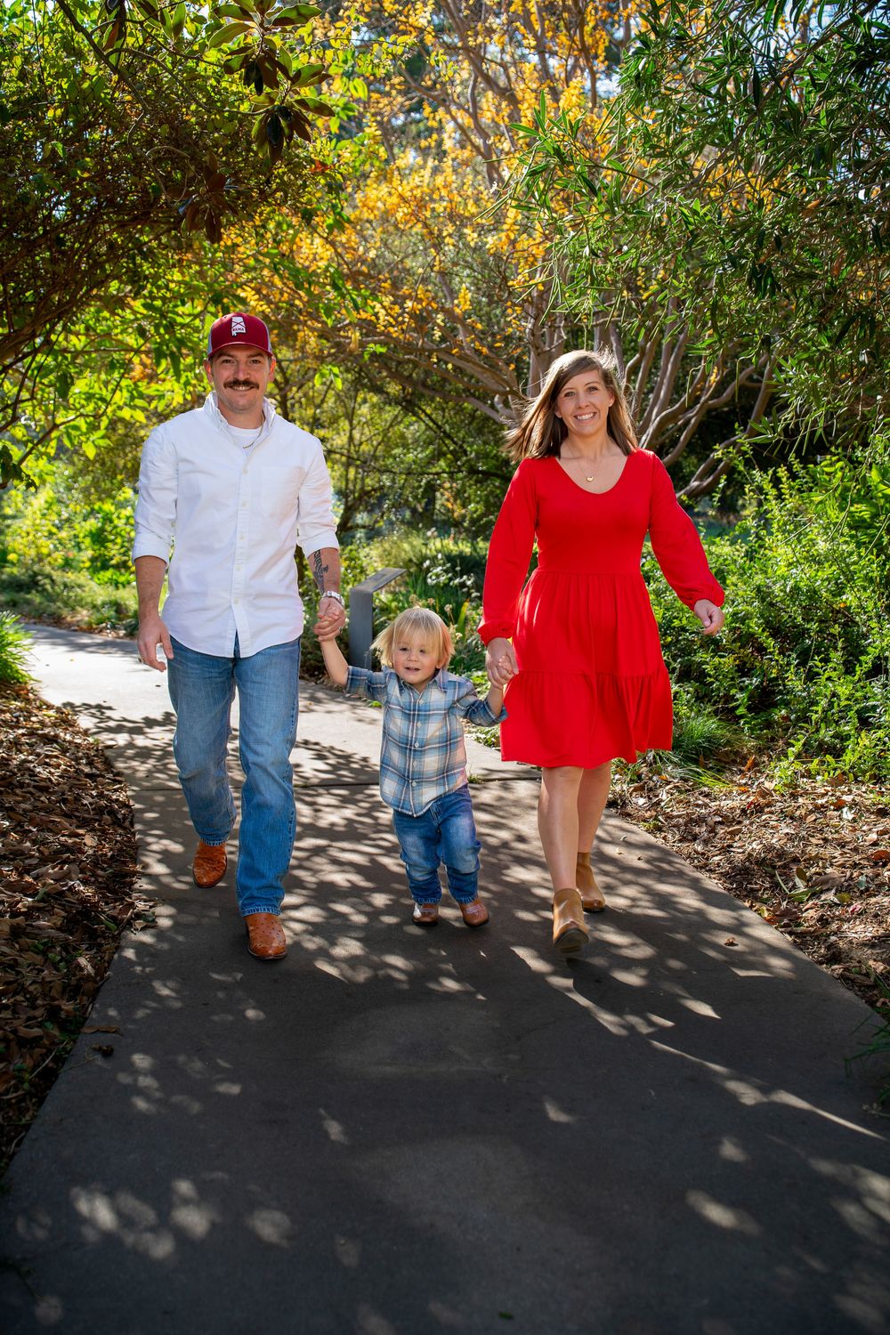 A happy family of three walks hand-in-hand along a tree-lined path in a lush green park.