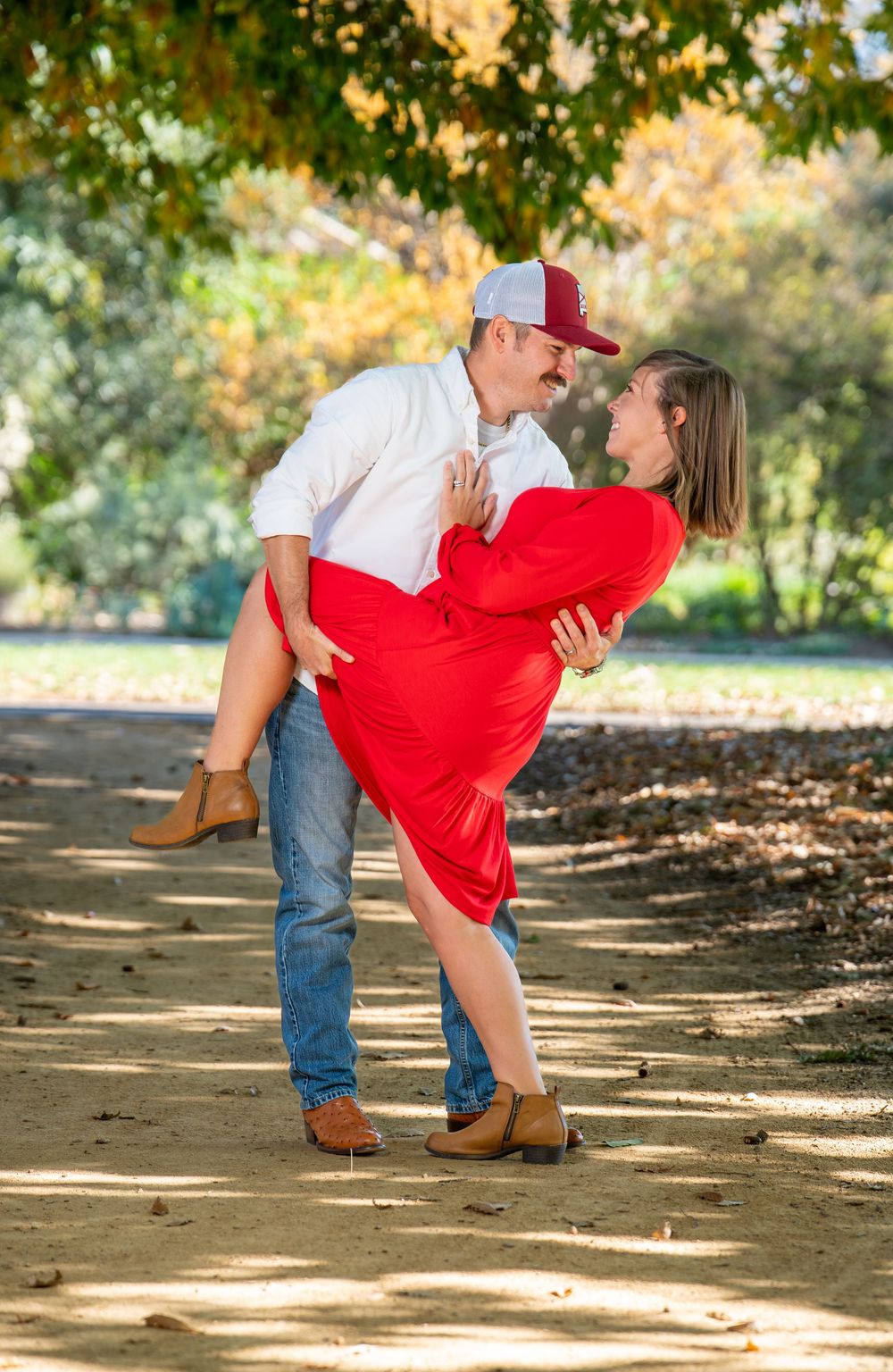 A couple shares a romantic dip embrace on a sunlit autumn park path under colorful foliage.