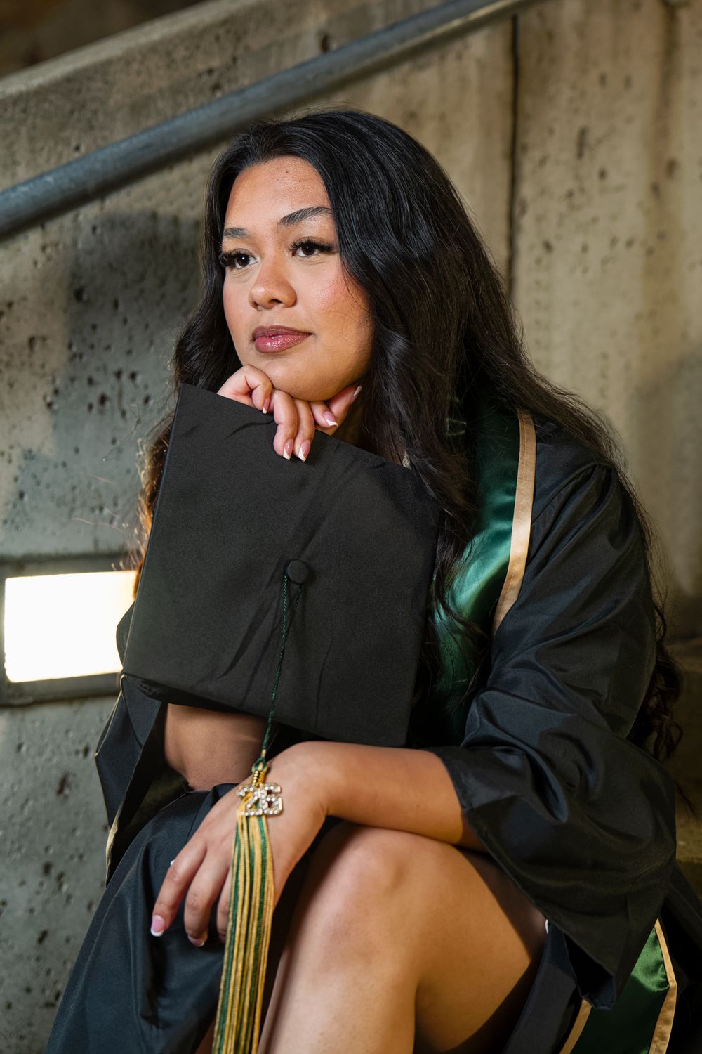A young woman in a black graduation gown and cap sits thoughtfully on concrete steps outdoors.