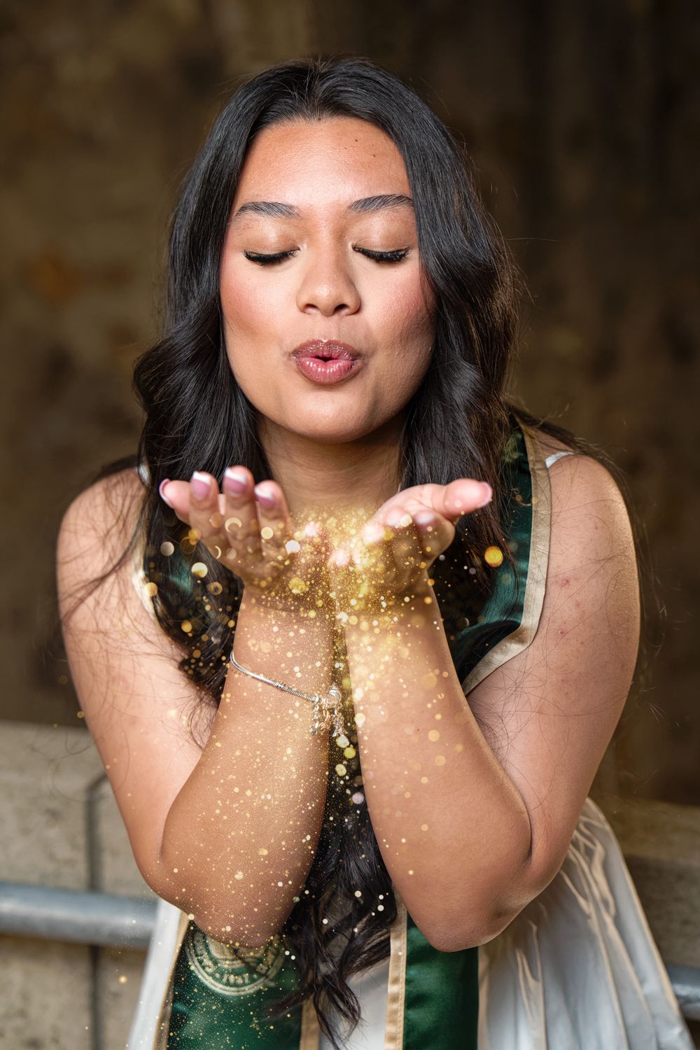 A young woman in graduation attire blows golden glitter from her hands with eyes closed.