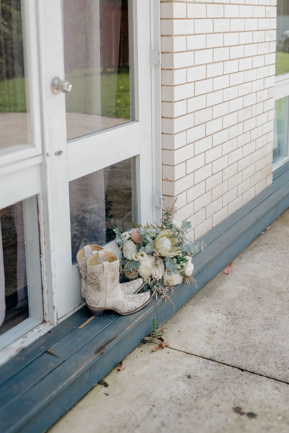 White cowboy boots and a white floral bouquet rest on a blue window sill beside a brick wall and glass door.