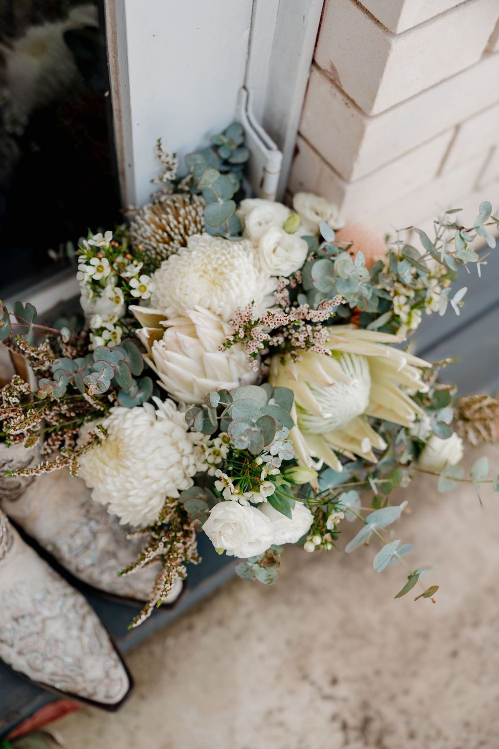 White wedding bouquet with protea, chrysanthemums, eucalyptus, and wax flowers beside lace bridal shoes on a doorstep.