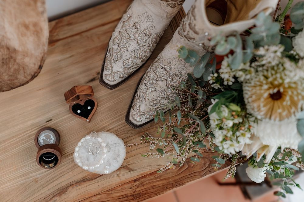 Rustic wedding flat lay with lace cowboy boots, floral bouquet, ring boxes, and garter on a wooden surface.