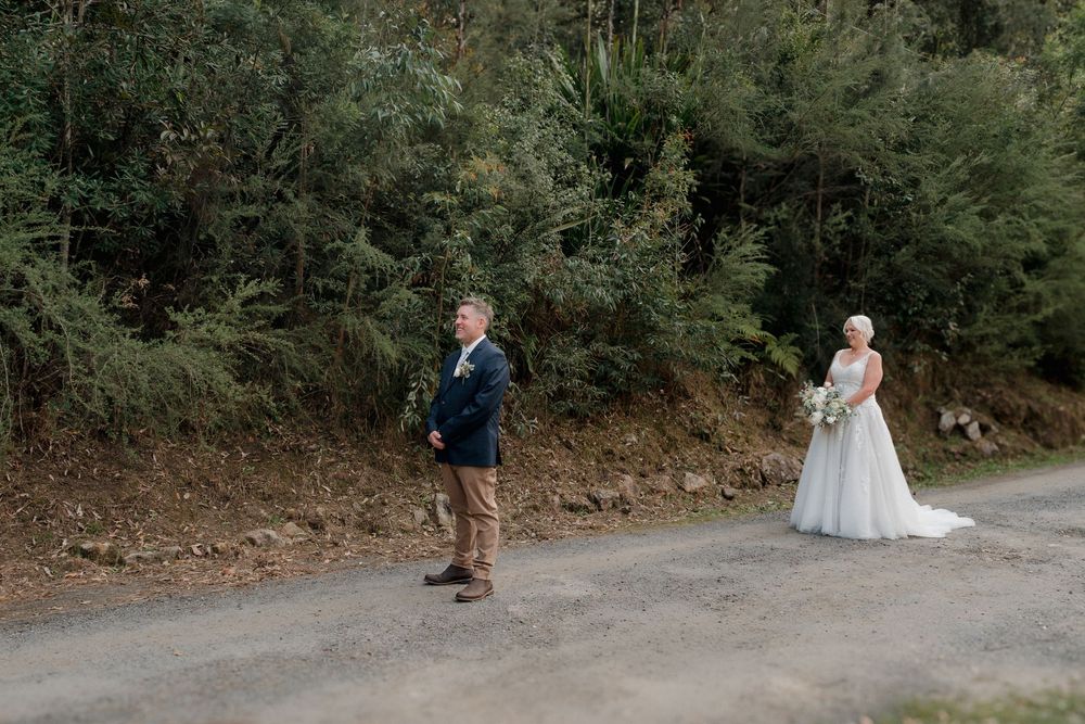 Bride in white gown holds bouquet while groom stands on a tree-lined path during outdoor wedding first look.