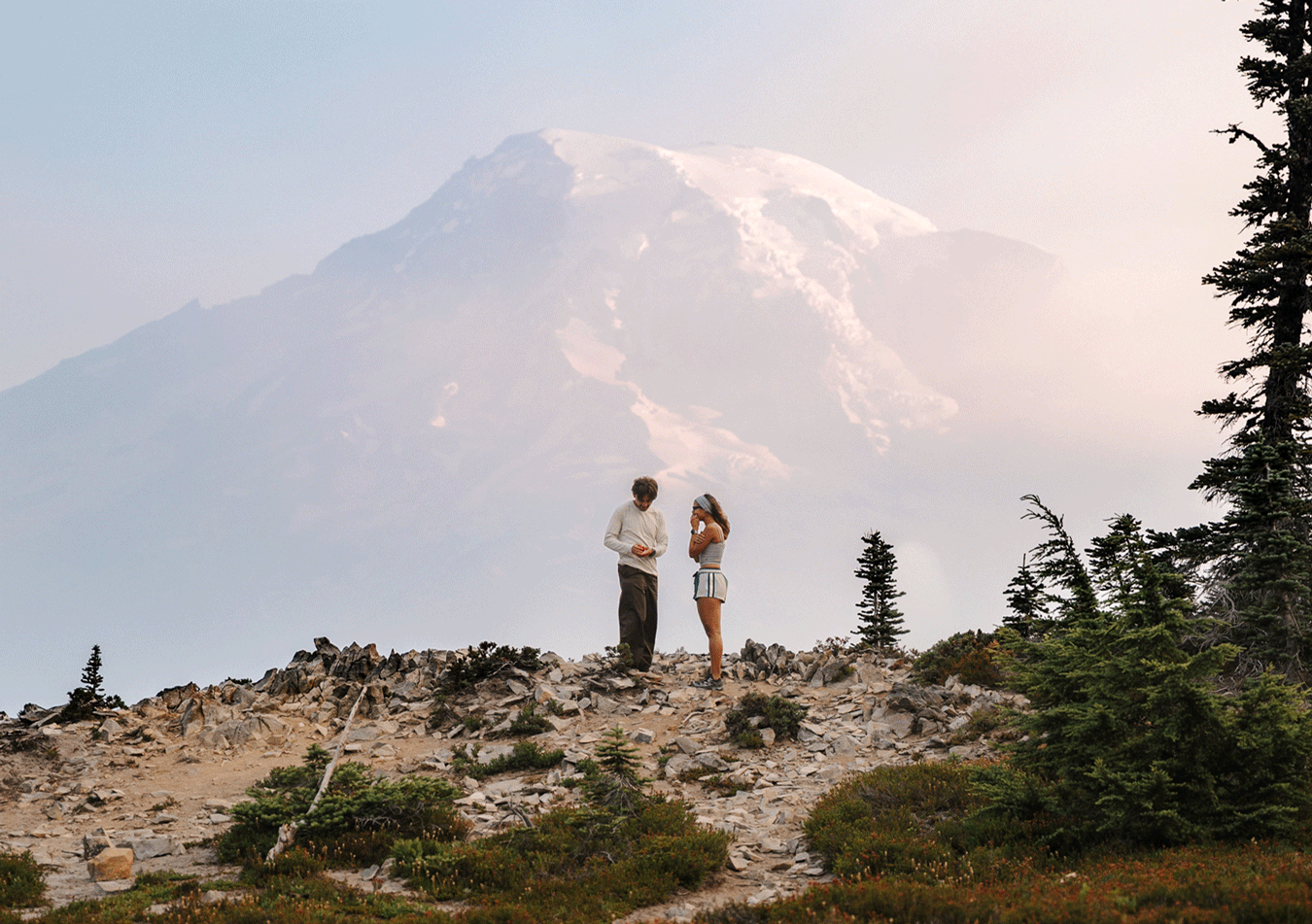Couple standing on rocky mountain ridge with misty snow-capped Mount Rainier in the background.