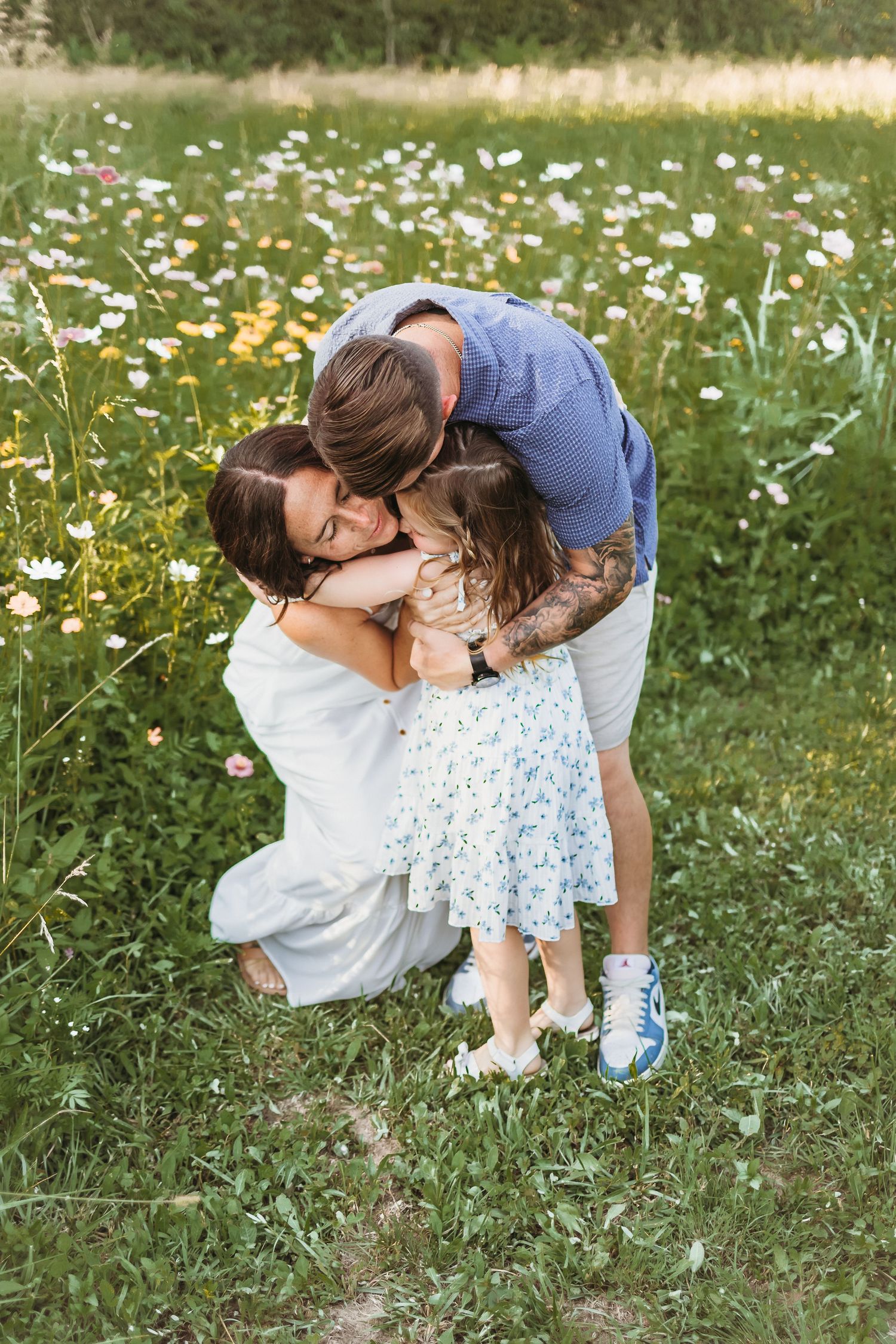 A family shares a tender embrace in a wildflower meadow, with parents hugging their young daughter outdoors.