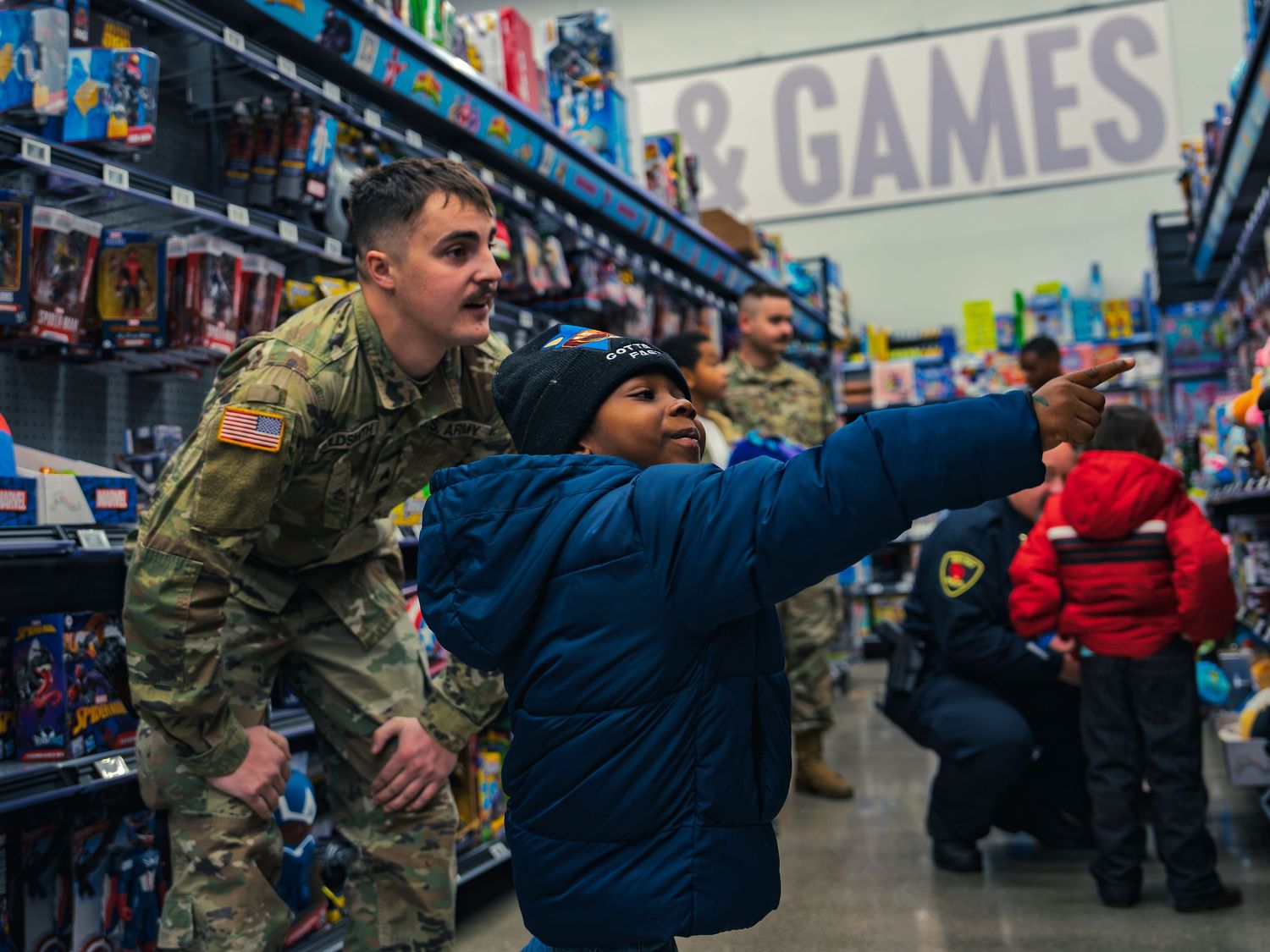 A child in a blue coat points down a toy aisle beside a National Guard soldier, with more soldiers and an officer shopping with kids behind.
