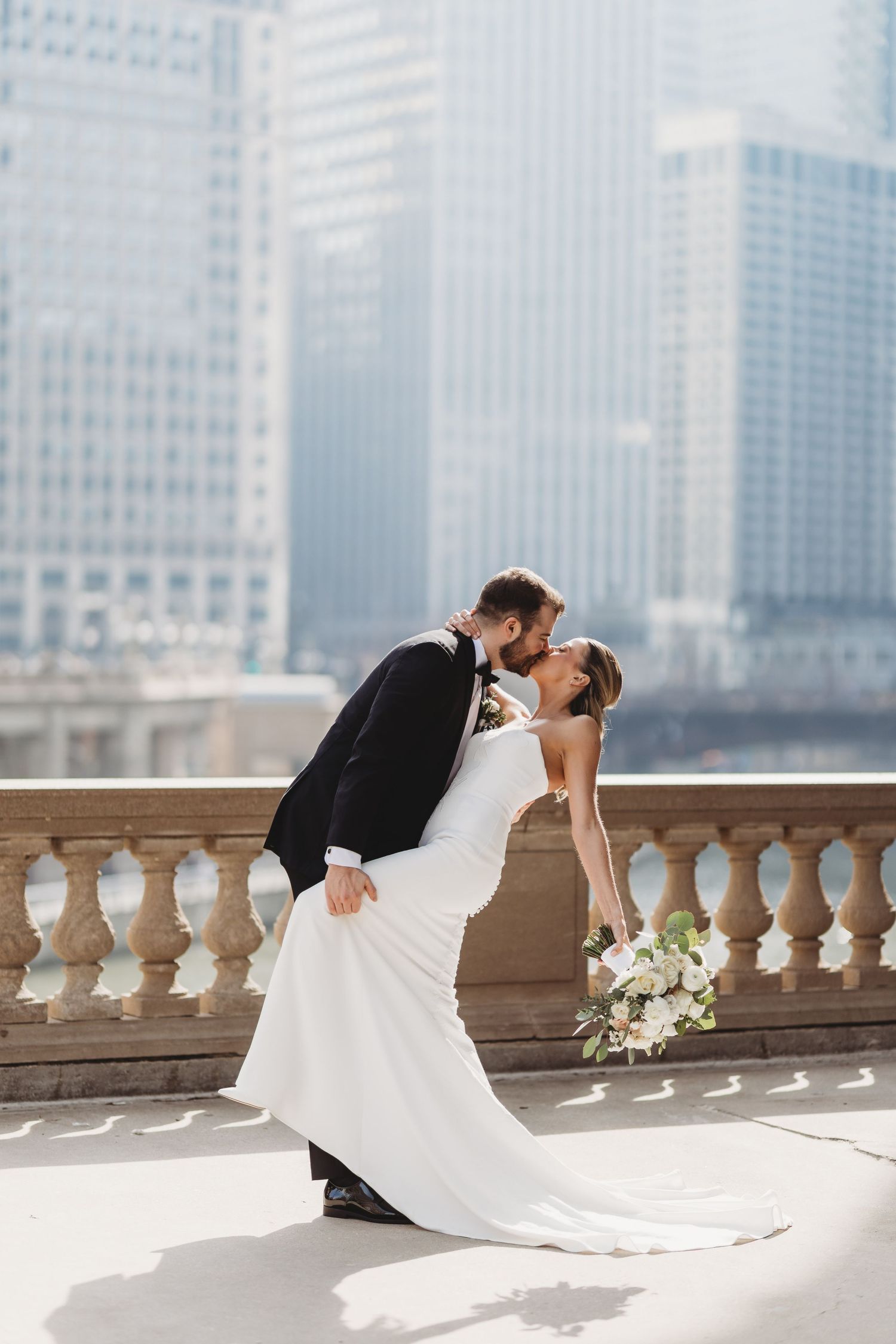 Bride and groom share a romantic dip kiss on a balcony with Chicago city skyline in the background.