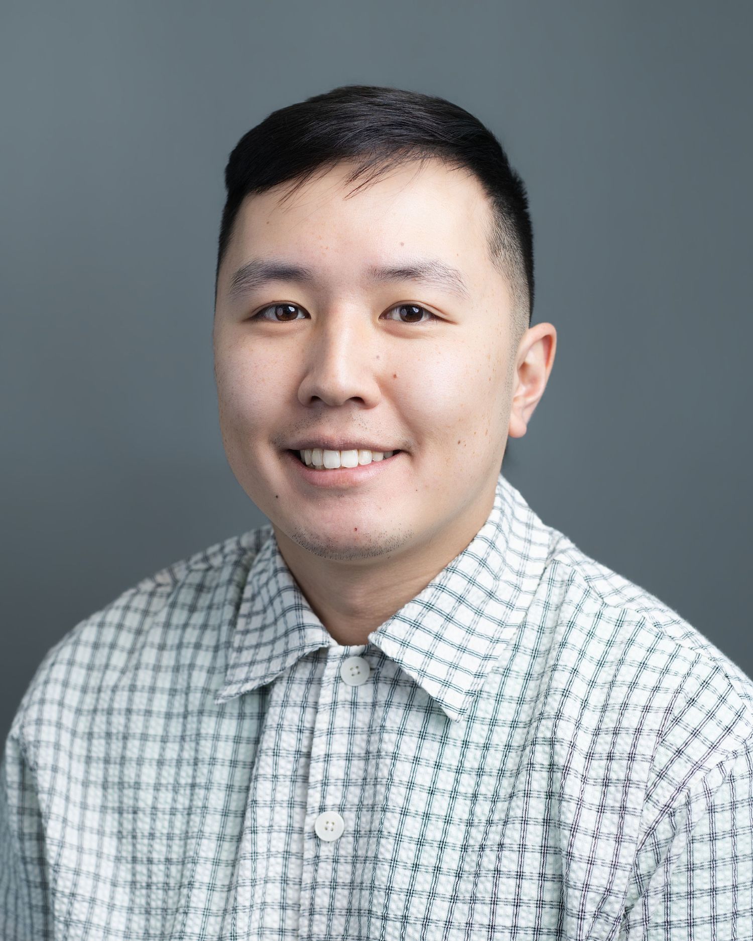 Professional headshot of a smiling young Asian man in a plaid shirt on a gray background.