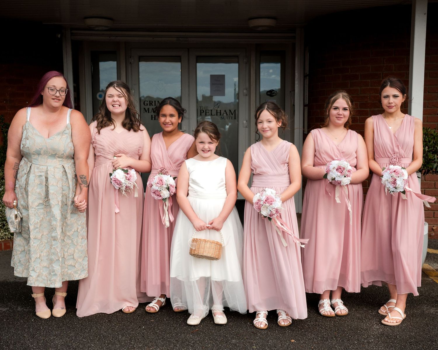 Bridesmaids in pink dresses and a flower girl in white pose together outside a venue holding pink bouquets.