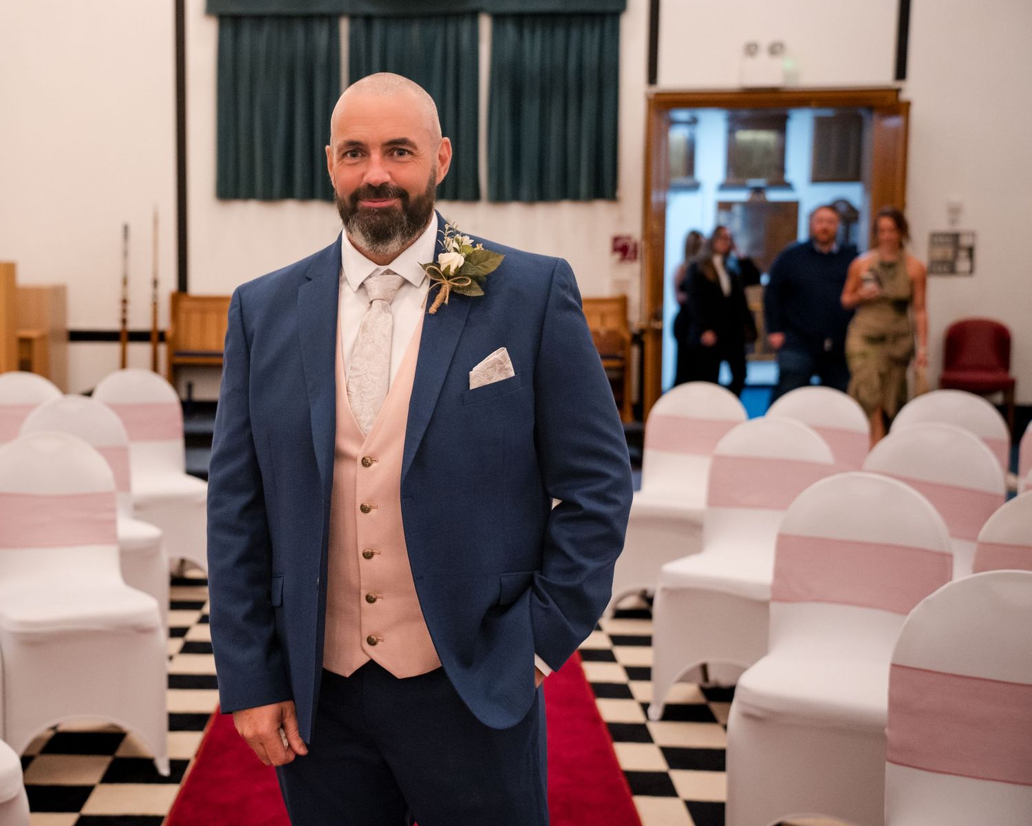 Groom in blue suit and champagne waistcoat with boutonniere stands in decorated wedding venue with pink chair covers.