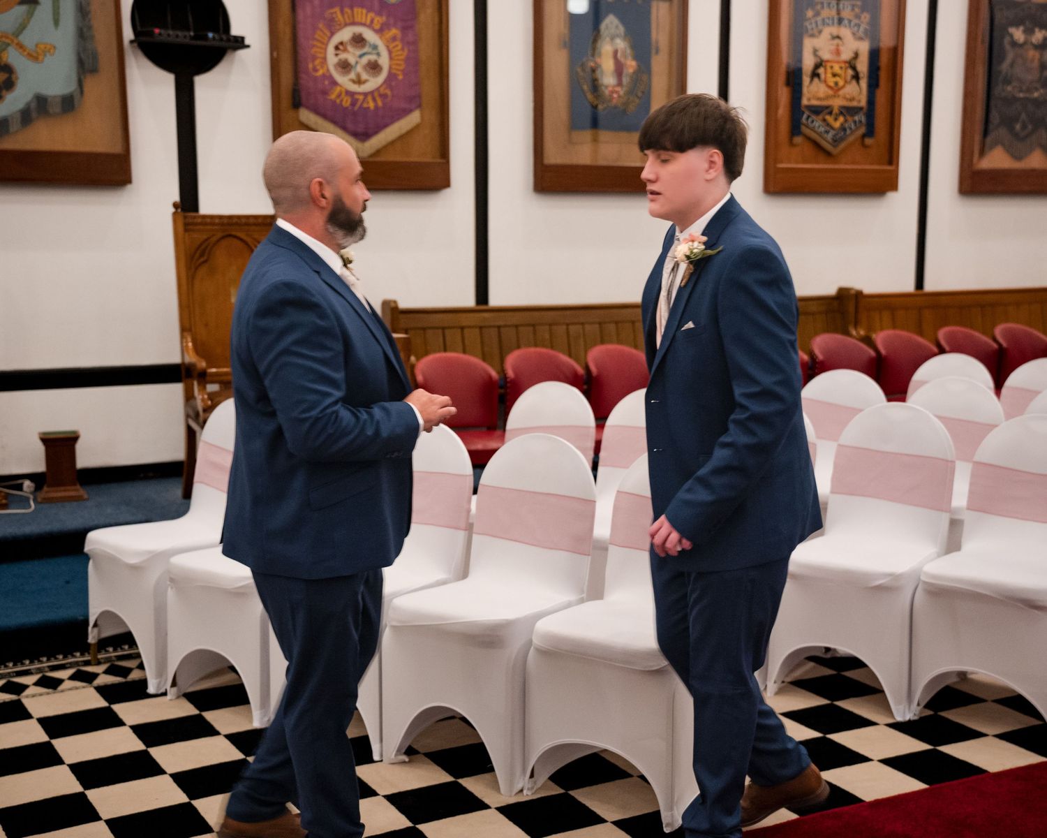 Two men in navy suits shake hands at a wedding venue with checkered floors and decorative banners.