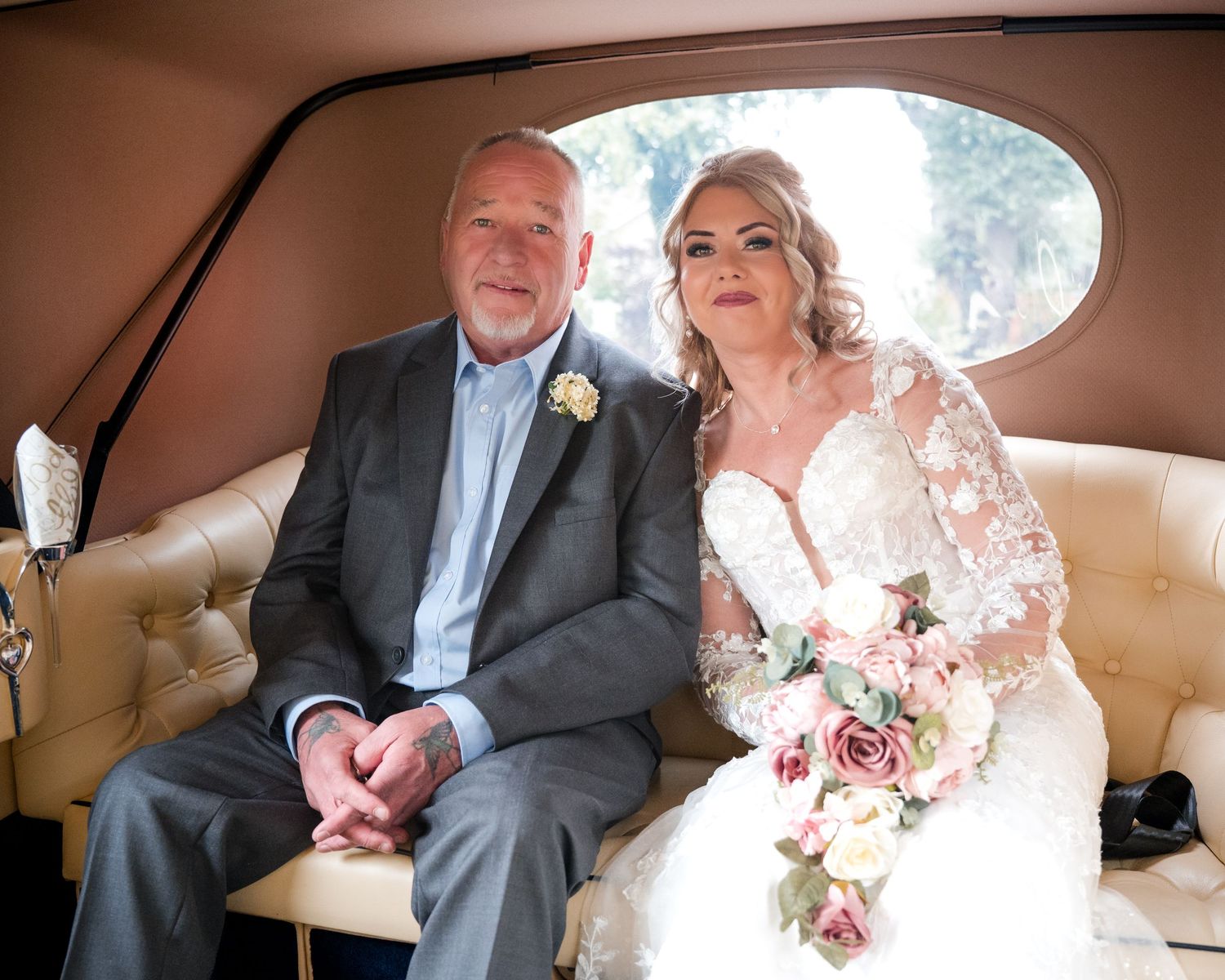 Bride and groom sitting together in a vintage car, bride holding pink bouquet in white lace gown.