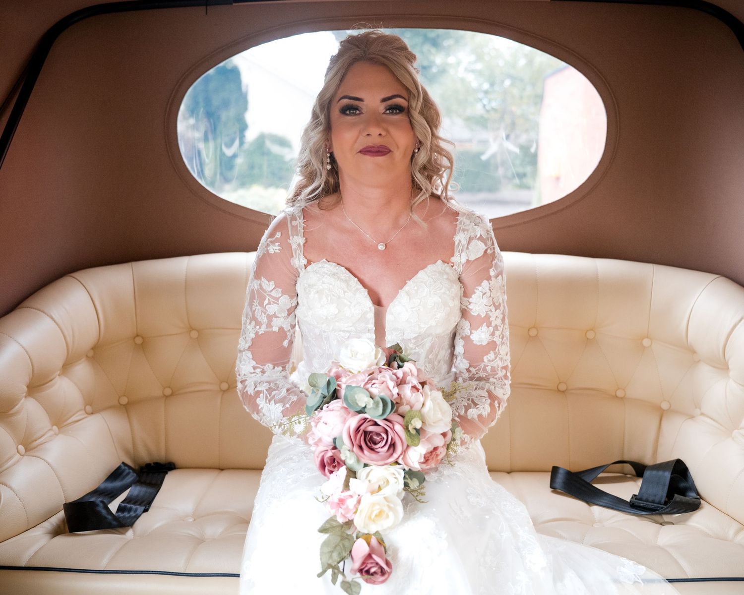 Bride in lace long-sleeve wedding gown holding pink rose bouquet, seated in cream vintage car interior.