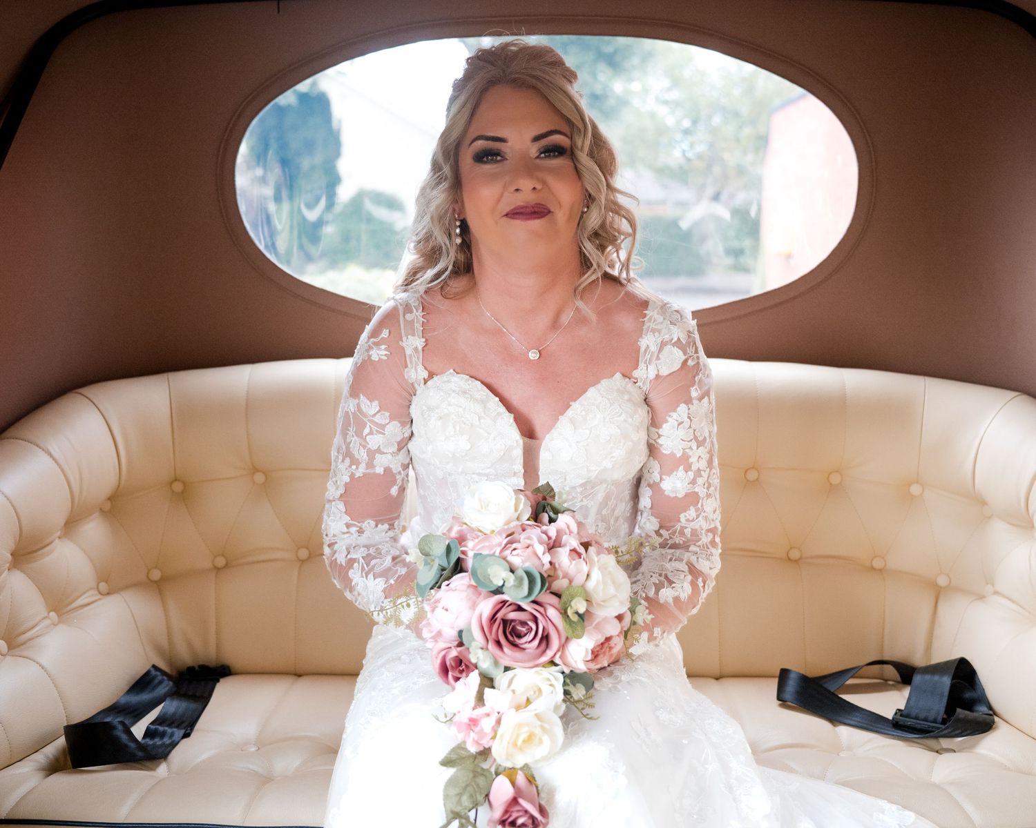 Bride in lace long-sleeve wedding gown holding floral bouquet seated in vintage cream leather car interior.