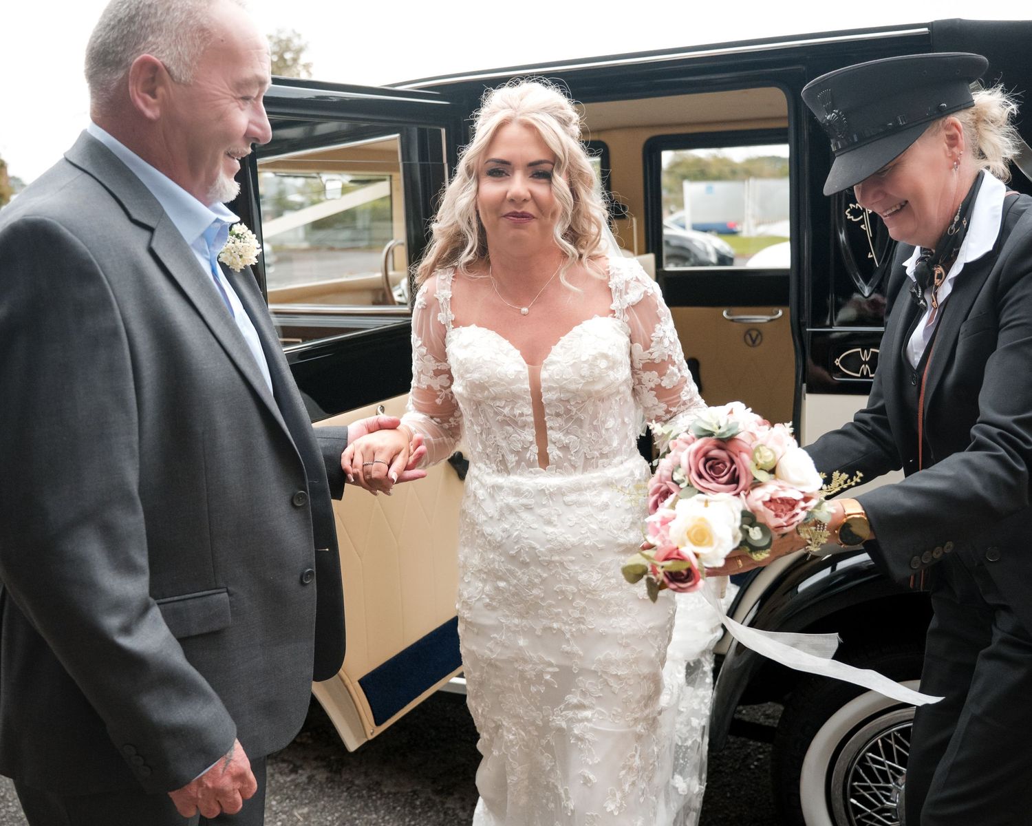 Bride in lace wedding dress holding bouquet beside vintage car with chauffeur and escort.