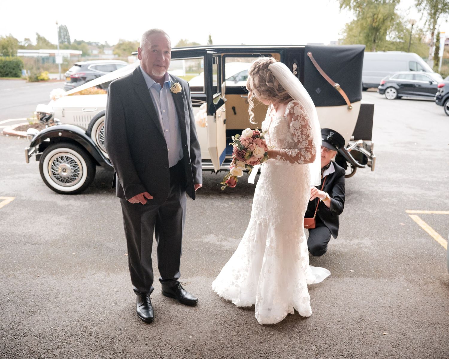 Bride in lace gown with bouquet stands beside suited man near vintage wedding car in parking lot.