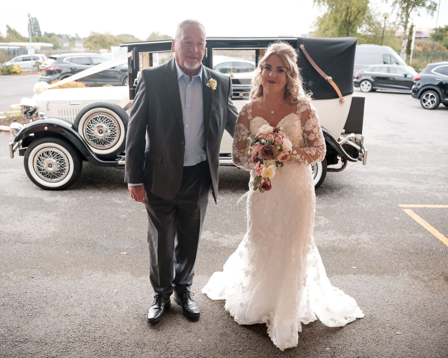 Bride in lace wedding dress holding bouquet stands with older man in suit beside vintage white wedding car.