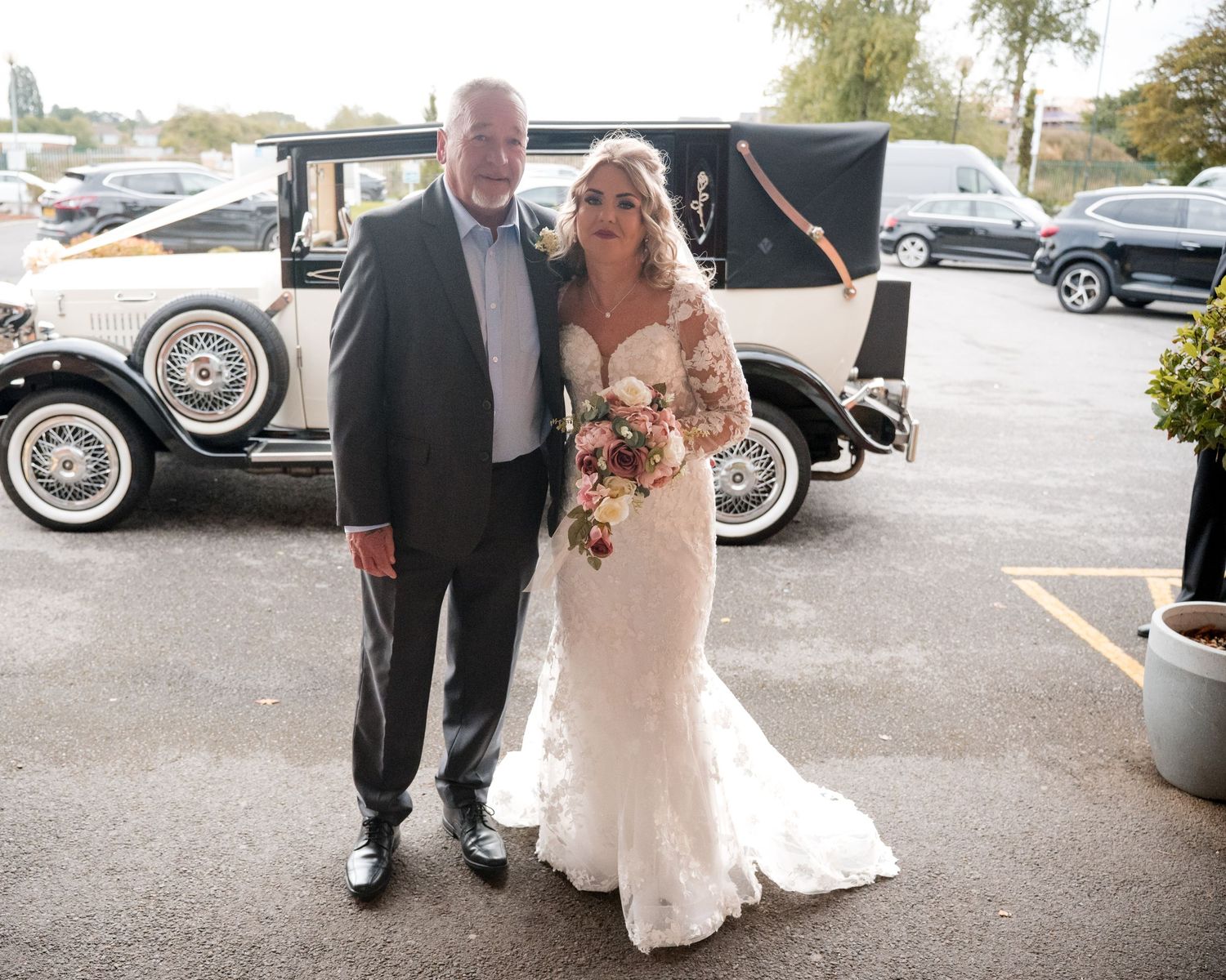 Bride in lace wedding gown holds bouquet beside older gentleman near vintage white wedding car in parking lot.