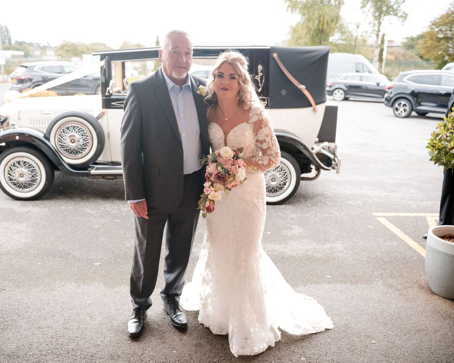 Bride in lace wedding gown holding bouquet poses with older gentleman beside vintage white classic car in parking lot.