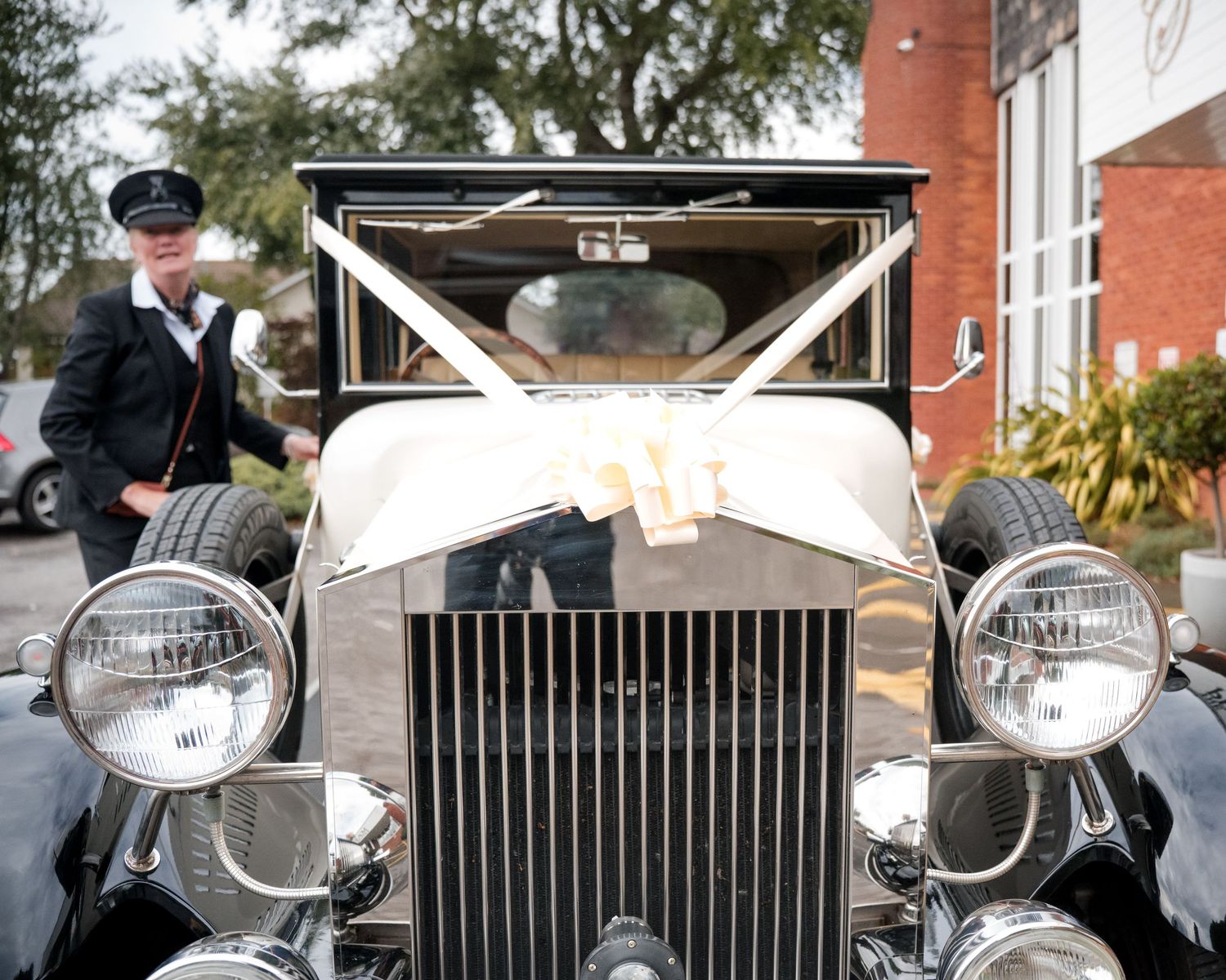 A uniformed chauffeur stands beside a vintage Rolls-Royce wedding car adorned with white ribbons outside a venue.