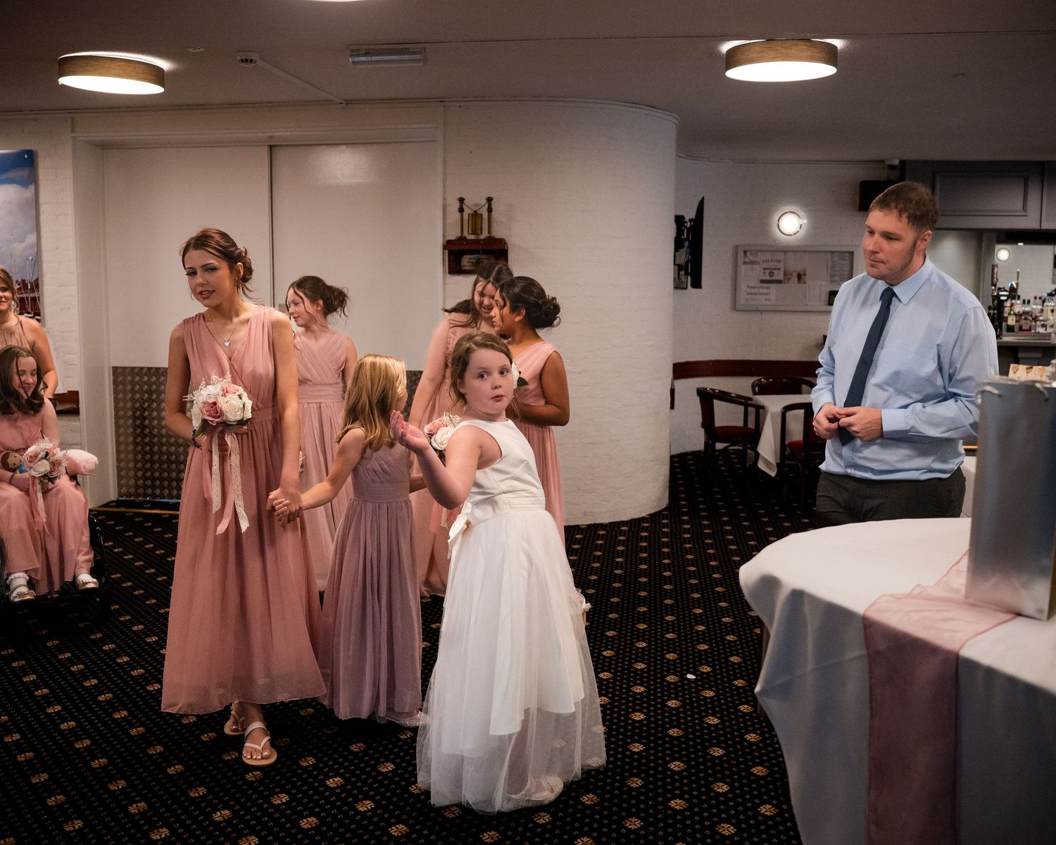 Bridesmaids in pink dresses with flower girls in white gather indoors at a wedding reception with a man in a blue shirt.