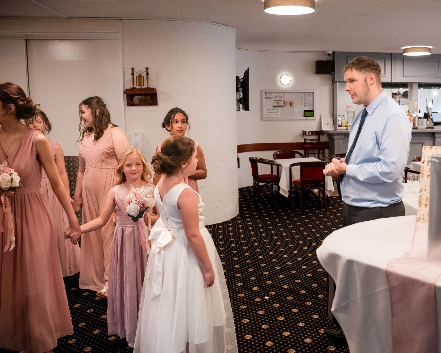 Wedding party with bridesmaids in dusty rose gowns and flower girls in white dresses gathered indoors near a decorated table.