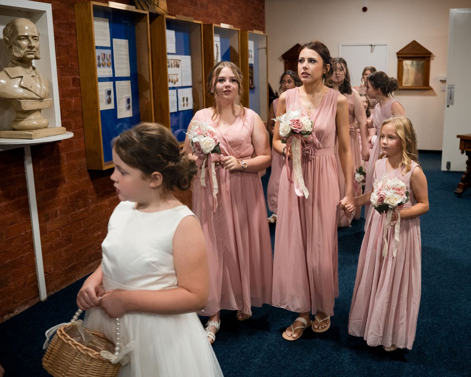 Bridesmaids in pink dresses holding bouquets walk with flower girls in white during a wedding ceremony procession.
