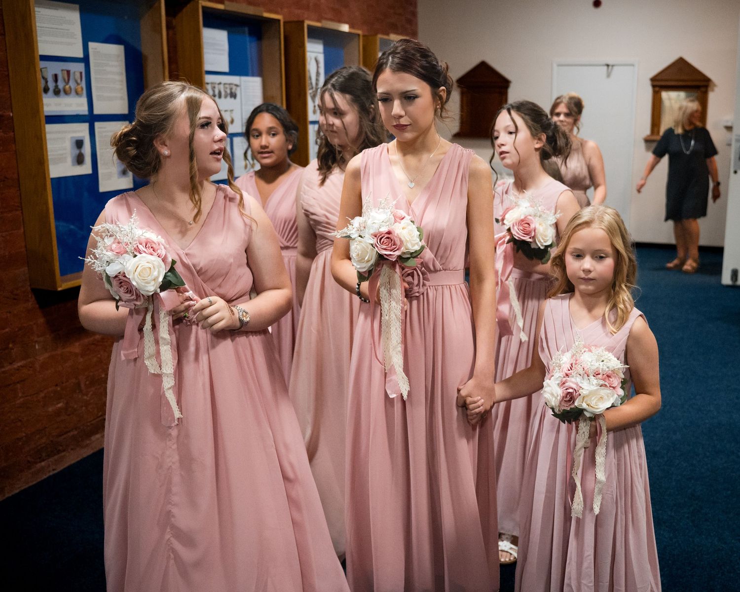 Bridesmaids in pink gowns holding white bouquets with a flower girl in a church hallway.