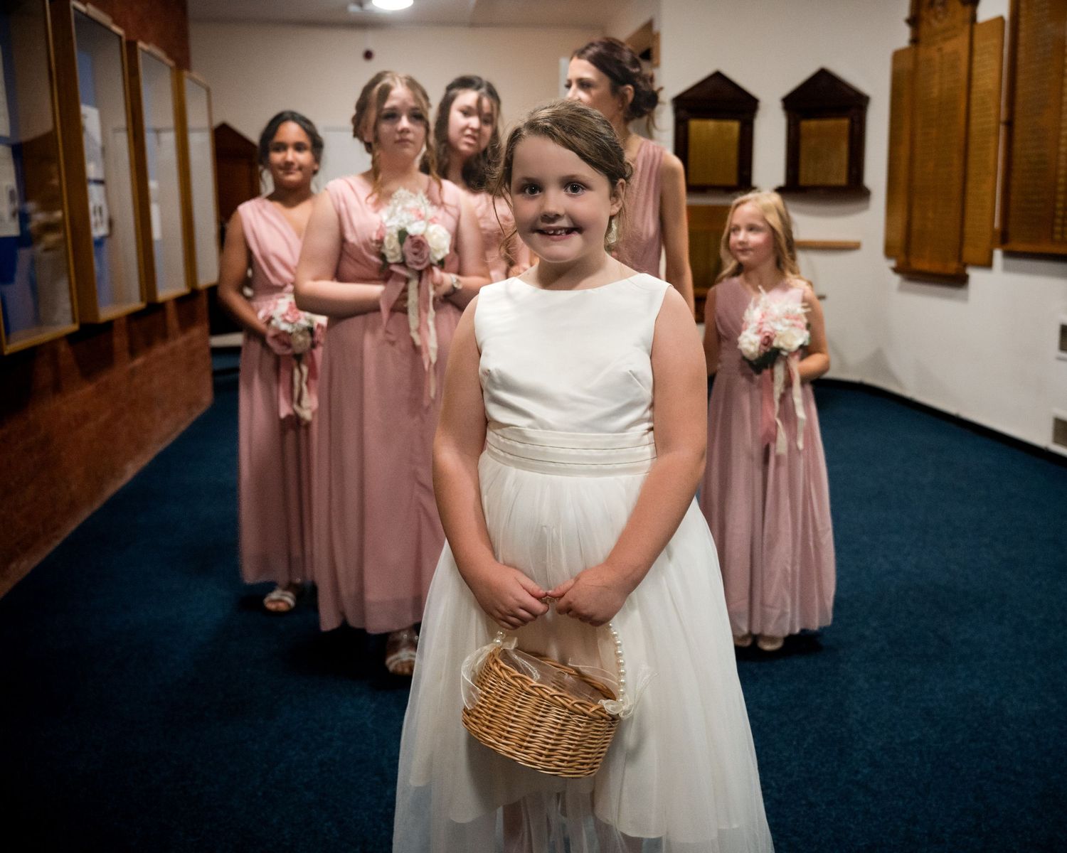 A young flower girl in a white dress holds a wicker basket, standing before bridesmaids in dusty pink gowns at a wedding.