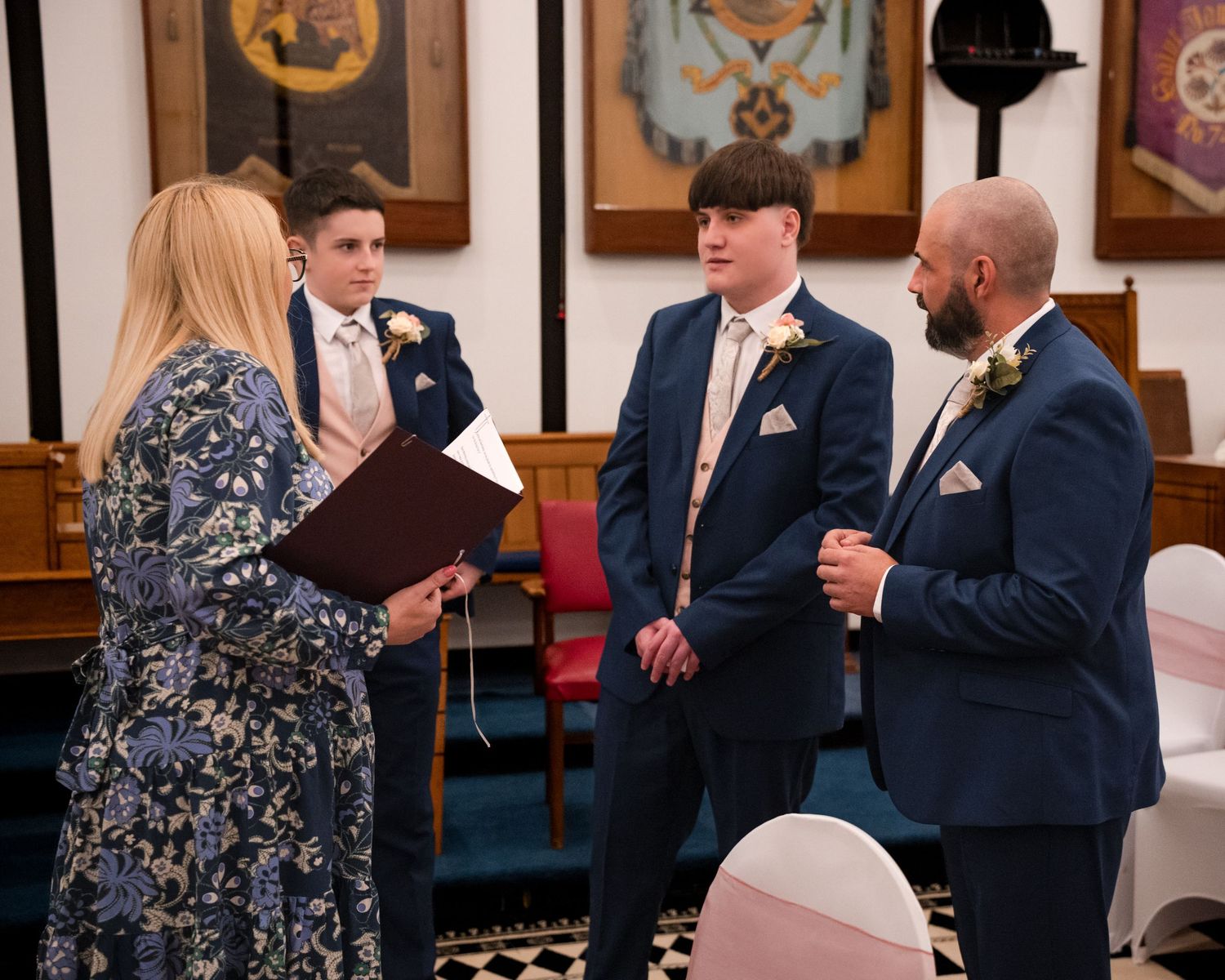 Three men in navy suits with boutonnieres stand before a woman officiant at an elegant wedding ceremony indoors.