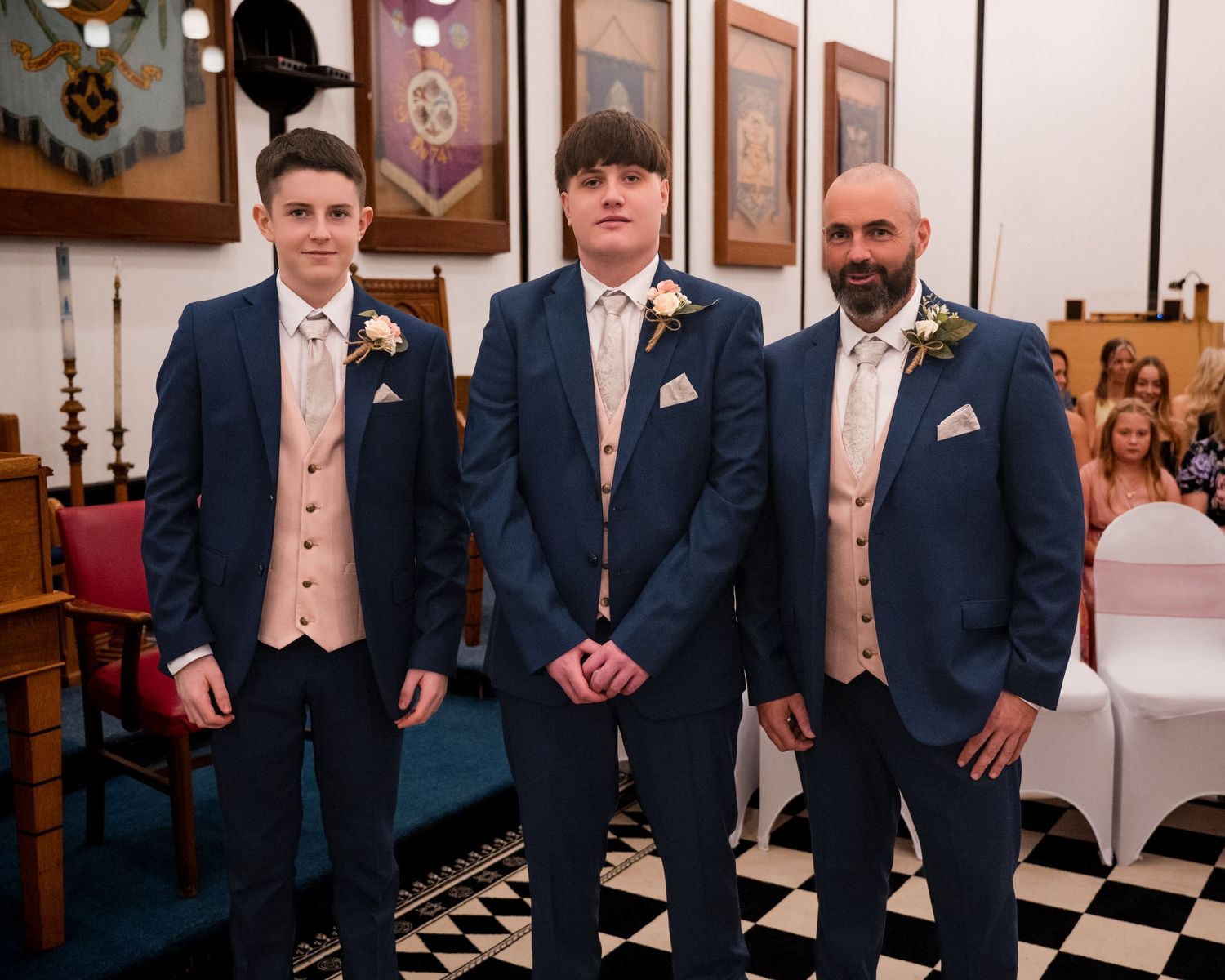 Three males in matching navy blue suits with champagne waistcoats pose together at a formal wedding ceremony.