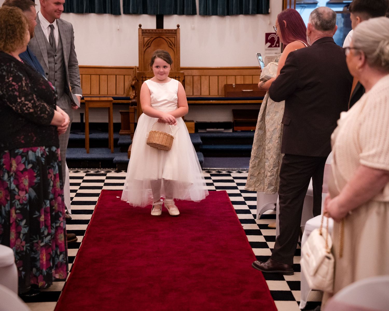 Young flower girl in white dress walks down red carpet aisle at wedding ceremony.