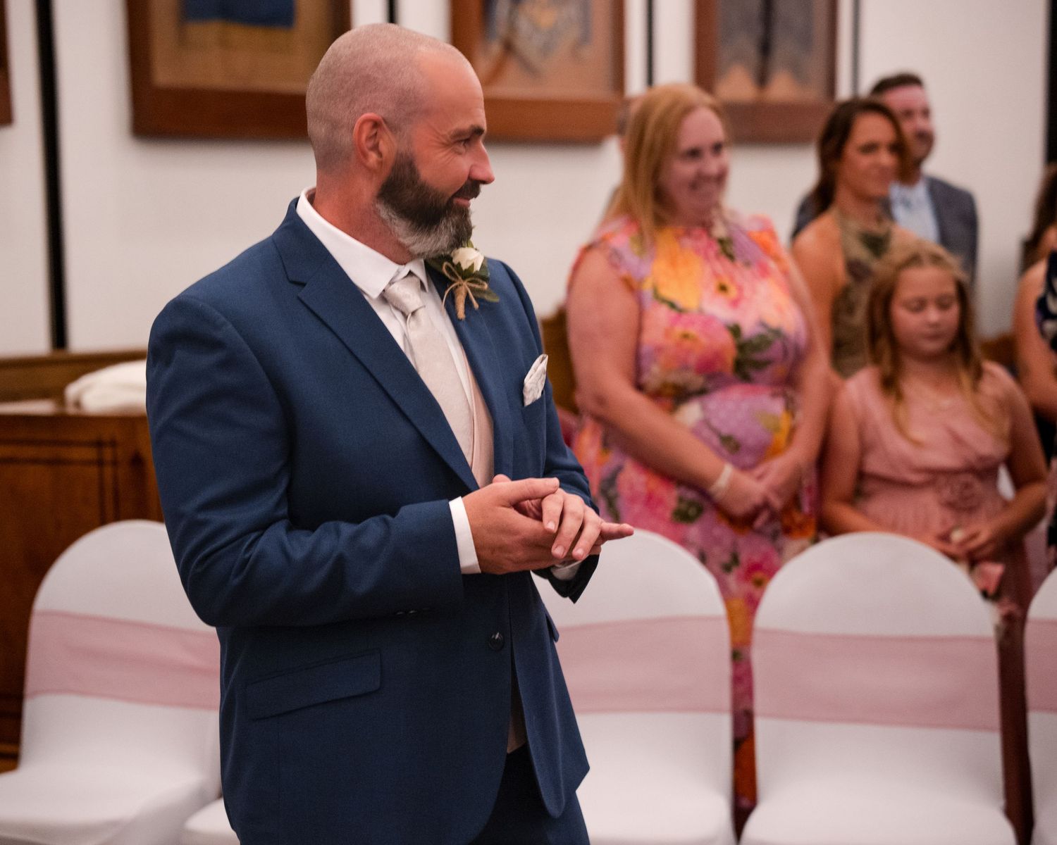 Groom in navy suit awaits bride at wedding ceremony, guests seated on white chairs with pink ribbon decor.