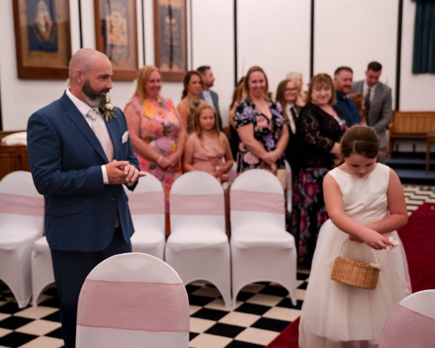 Groom in blue suit watches flower girl scatter petals at wedding ceremony.