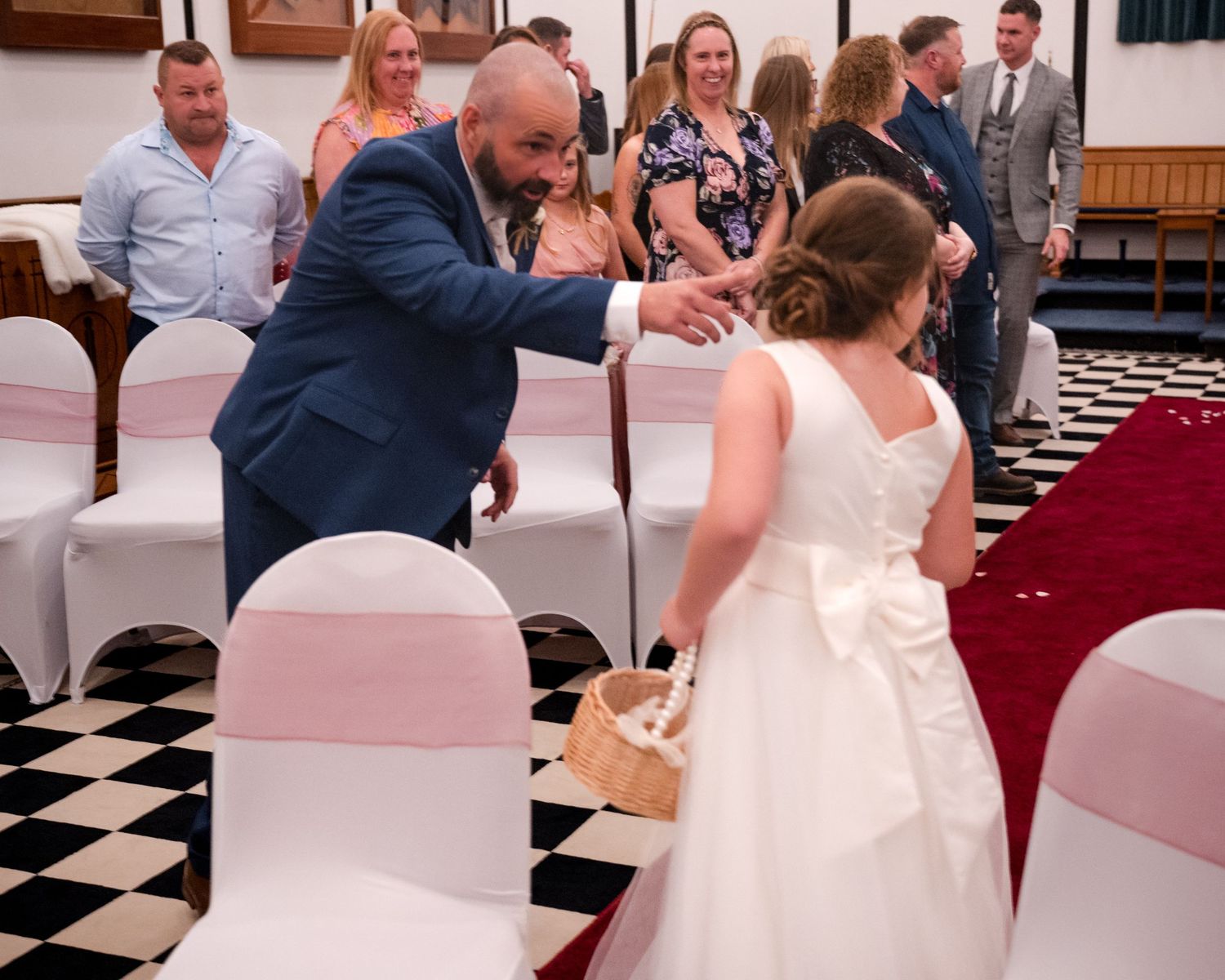Man in blue suit greeting flower girl in white dress at a wedding ceremony with guests seated on white chairs.