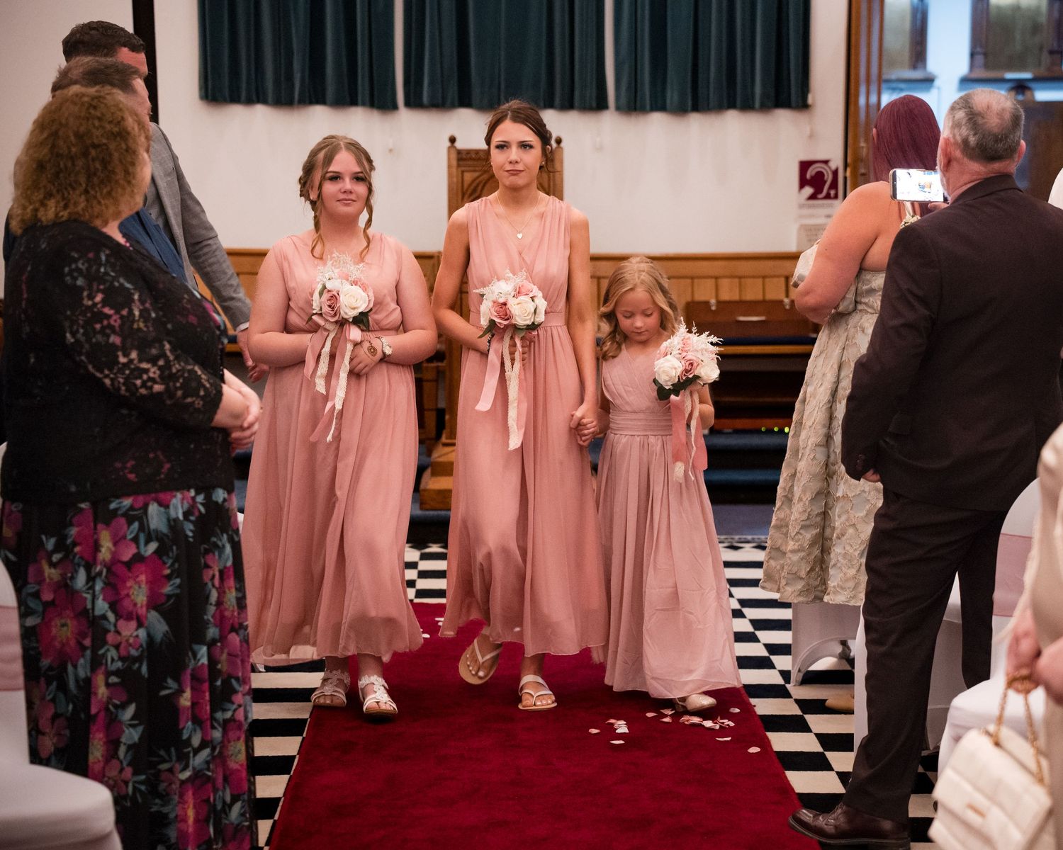 Three bridesmaids in pink dresses walk down a red carpet aisle in a church during a wedding ceremony.