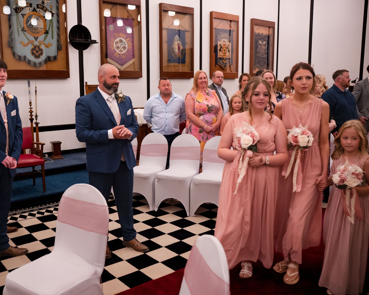 Bridesmaids in blush pink gowns holding bouquets walk down the aisle in a decorated wedding ceremony venue.