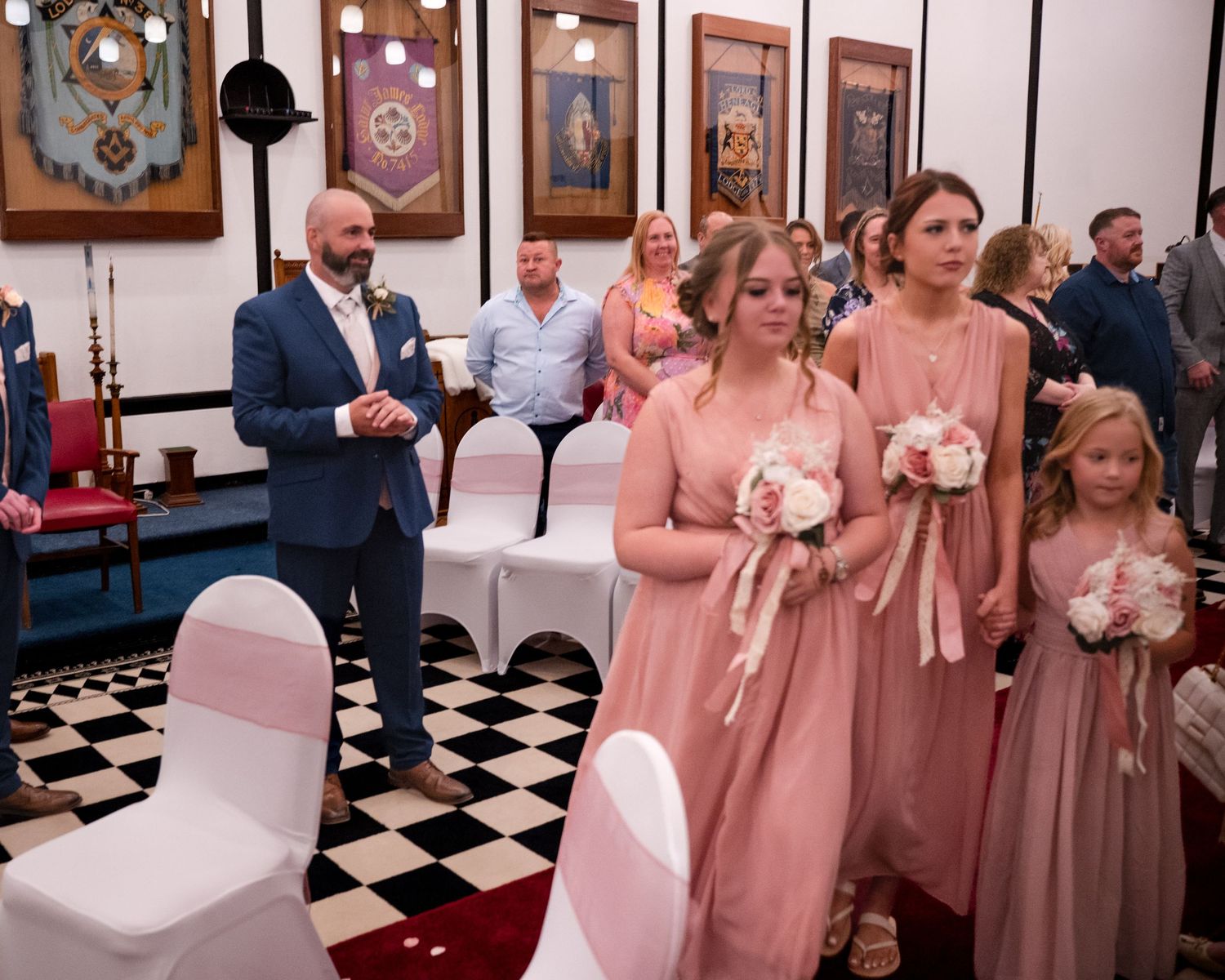 Bridesmaids in pink dresses walk down the aisle at an elegant wedding ceremony in a decorated hall.